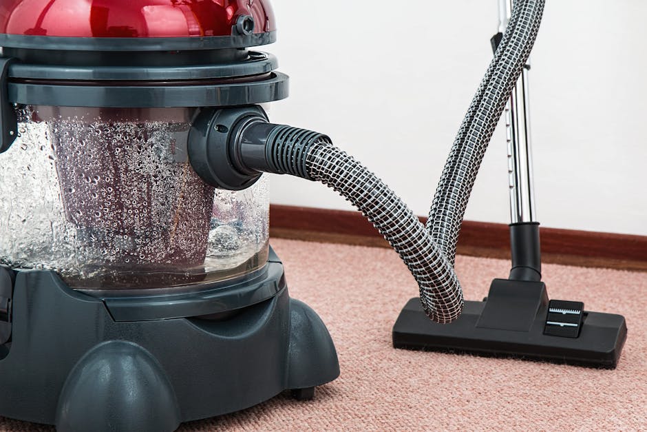 Close-up of a modern vacuum cleaner with a transparent dust container filled with particles, connected to a flexible hose and upright extension, positioned on a light pink carpet in a residential room. The vacuum cleaner features a red and black body, and the scene is well-lit with natural or artificial light, highlighting the clean and tidy appearance of the carpet surface. The background includes a white wall and a wooden baseboard, emphasizing an indoor domestic setting. The image reflects professional surface cleaning, showcasing equipment used by Carpet Cleaning Surrey for deep cleaning and sanitisation of carpets in residential homes, such as those on London Road in Guildford GU1.