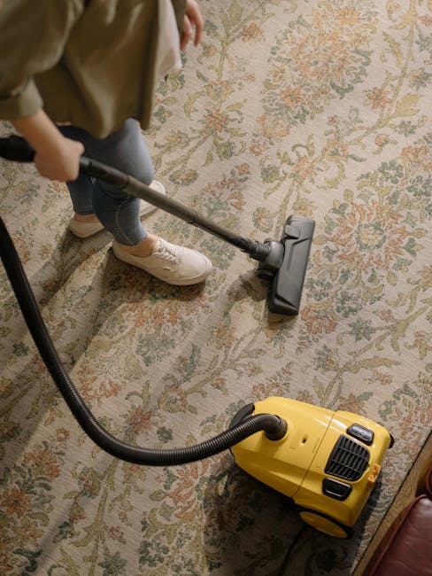 A person, wearing casual clothing including beige sneakers and blue jeans, is vacuuming a patterned Oriental-style carpet in a residential living room. The carpet has intricate floral designs in shades of beige, orange, and green. The individual is using a yellow and black vacuum cleaner with a long metal wand and a wide brush head to perform deep surface cleaning. The room is well-lit, highlighting the carpet's clean and well-maintained condition. The vacuum cleaner is placed on the floor, and the person’s hand is visible guiding the cleaner. This scene exemplifies professional domestic cleaning, focusing on carpet surface cleaning to maintain hygiene and appearance, representative of services offered by Carpet Cleaning Surrey, as referenced in the Guildford GU1 carpet cleaning guide for London Road homes, Surrey.