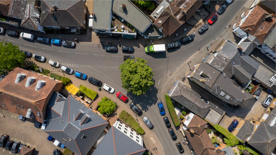 An aerial view of a small town street in Walton on Thames, showing a curved road lined with parked cars in various colors, including blue, red, black, and white. The street is bordered by residential 