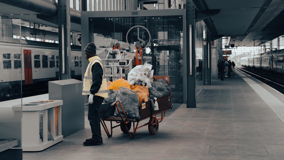 A cleaner in a high-visibility vest and knit cap stands next to a luggage trolley loaded with trash bags and debris at Woking station platform. The platform features clean, tiled surfaces with a smooth, glossy finish, illuminated by natural daylight. In the background, there are glass walls, train tracks, and parked trains, with minimal dust or dirt visible, indicating a maintained environment. The scene emphasizes routine surface cleaning and sanitation practices typical of commercial and transportation cleaning services, as provided by Carpet Cleaning Surrey, in alignment with the guide for commuters at Woking station.