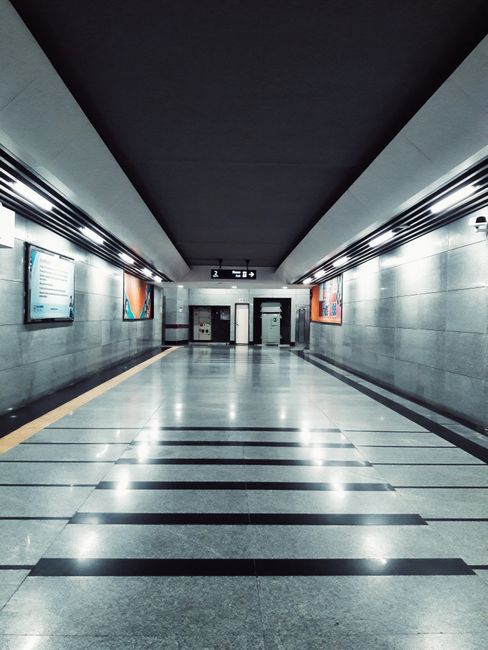 A clean, modern underground corridor at Woking station with polished, reflective marble or stone flooring featuring dark linear accents. The smooth, light gray walls are illuminated by evenly spaced, bright LED lighting strips running along the ceiling's edges. Three digital information screens are mounted on the left wall, displaying schedules or advertisements, while on the right wall, there are framed images enhancing the space's appearance. At the far end, two elevator doors are centered, with clear signage indicating their function. The overall scene portrays a well-maintained, hygienic environment, indicative of thorough deep cleaning and surface maintenance by Carpet Cleaning Surrey to ensure a pristine, safe area for commuters.